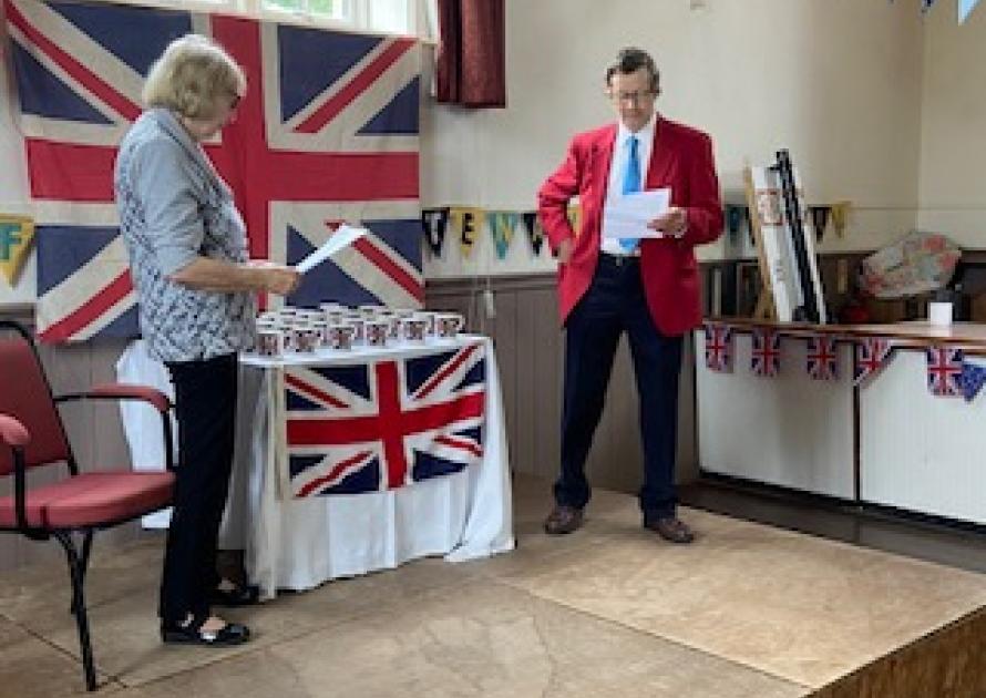 2 parishioners on stage in front of a Union Jack and table with commorative mugs
